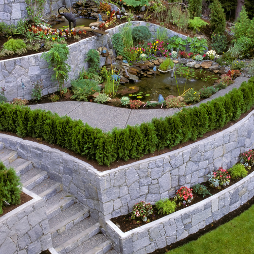 Stone terraced garden with a pond, waterfall, and various plants and flowers.