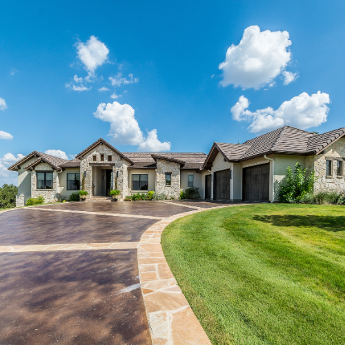 Elegant stone house with a long driveway and manicured lawn under a blue sky.