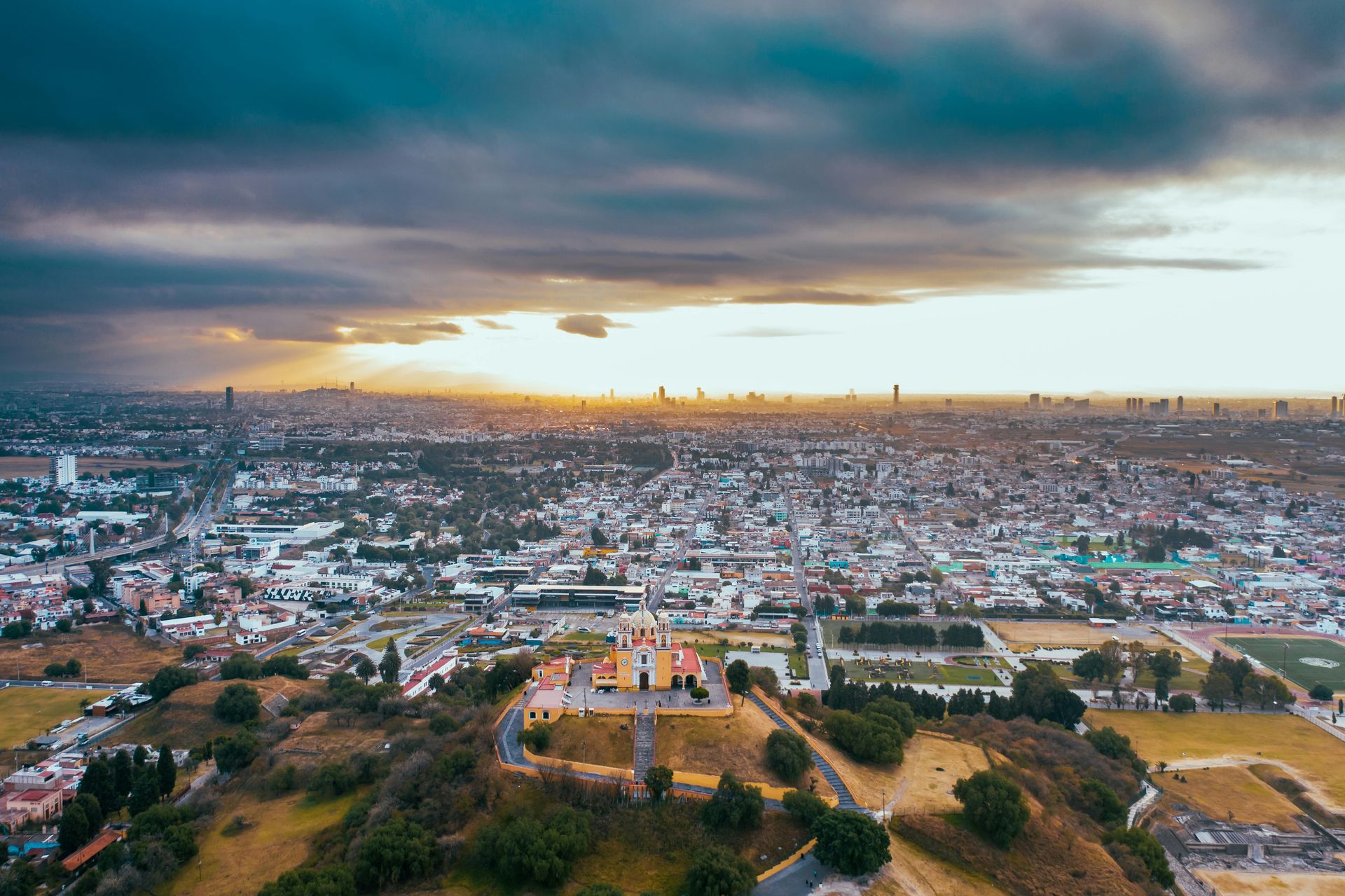 Cityscape with a hilltop church, sunset illuminating buildings under a cloudy sky.