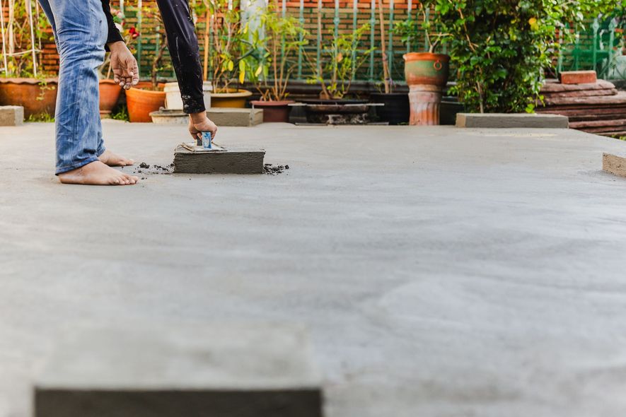 Person smoothing wet concrete with a trowel outdoors, feet bare, surrounded by plants.