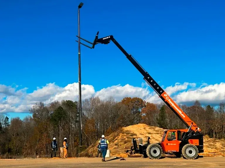 Orange forklift lifting a tall metal pole; two workers observe. Blue sky, trees in background.