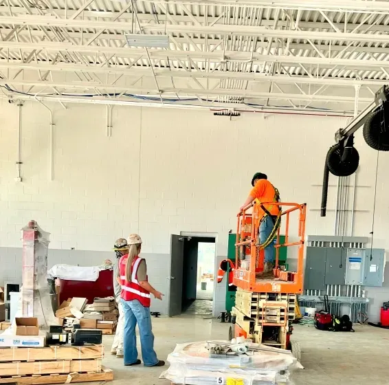 Construction workers in an open space; one on a lift, others standing, inspecting. Building materials scattered.