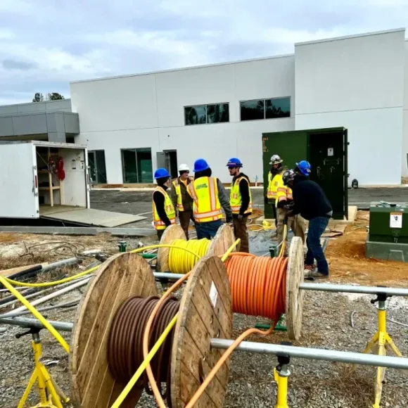 Construction workers in hard hats and vests installing power cables near a white building.