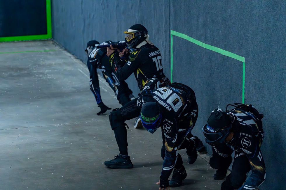 Paintball players in black and gold gear aiming guns behind a wall, indoors.