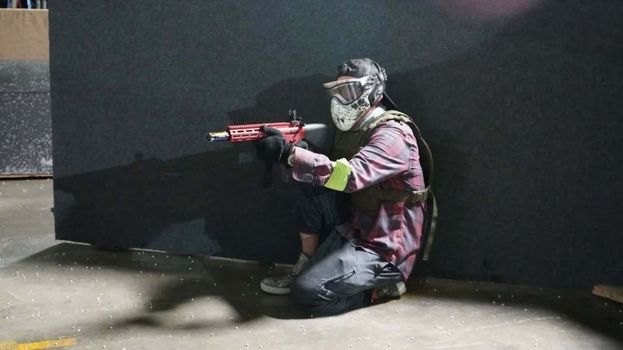 Person kneels, aiming a red paintball gun, wearing a mask and camo clothing, against a dark wall indoors.