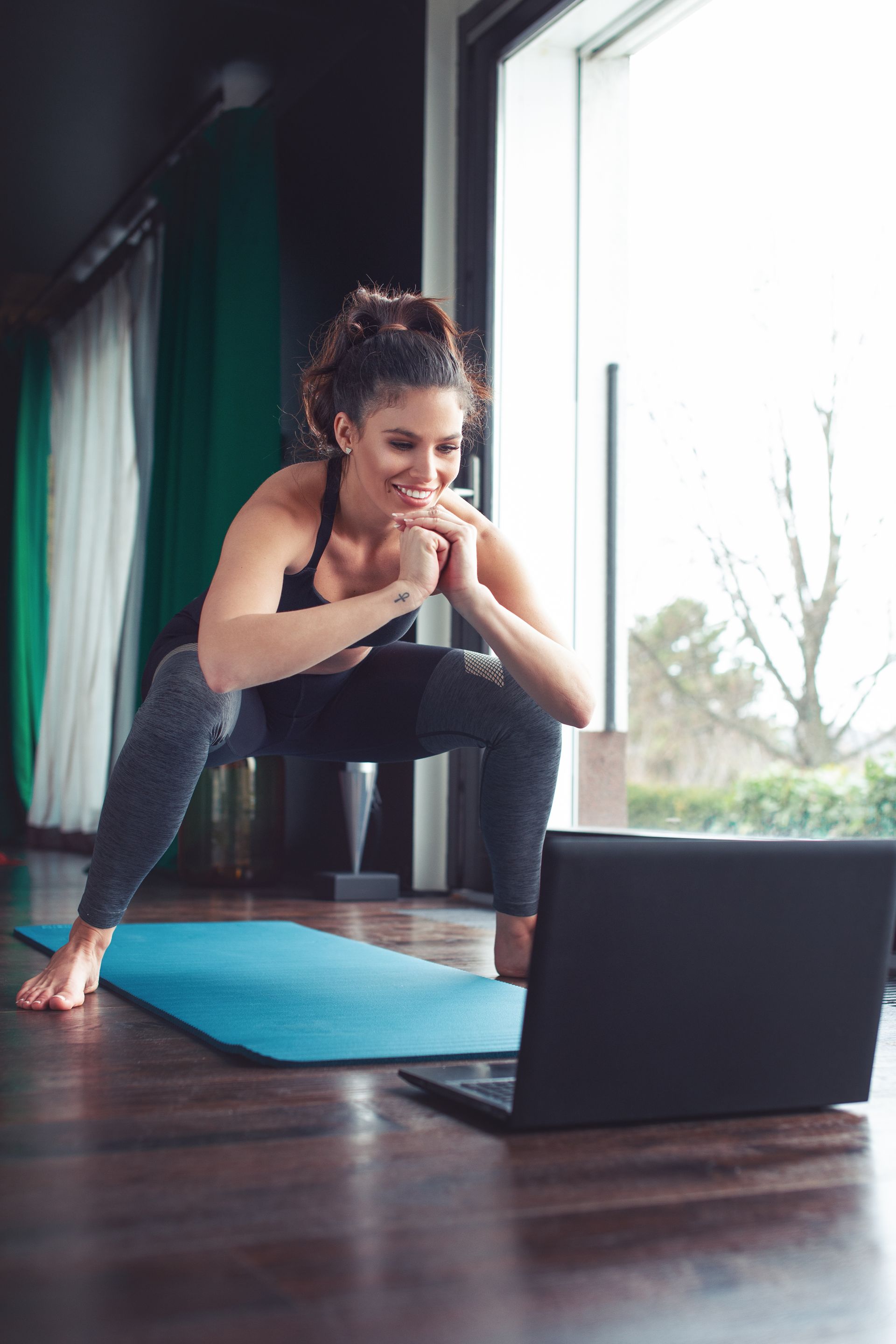 A woman is squatting on a yoga mat while looking at a laptop.