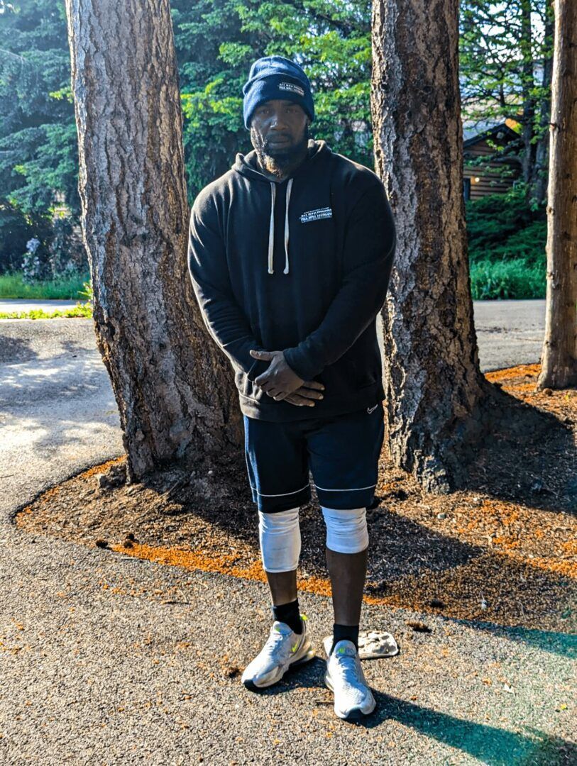 A man in a black hoodie and shorts is standing in front of trees.