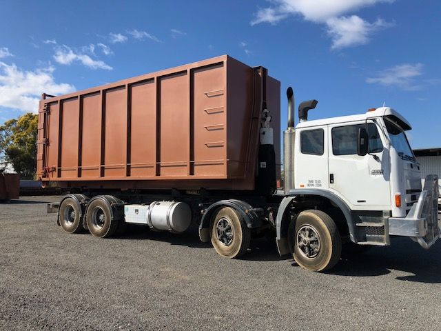 A brown industrial dumpster on a white truck, parked on a gravel lot under a blue sky.