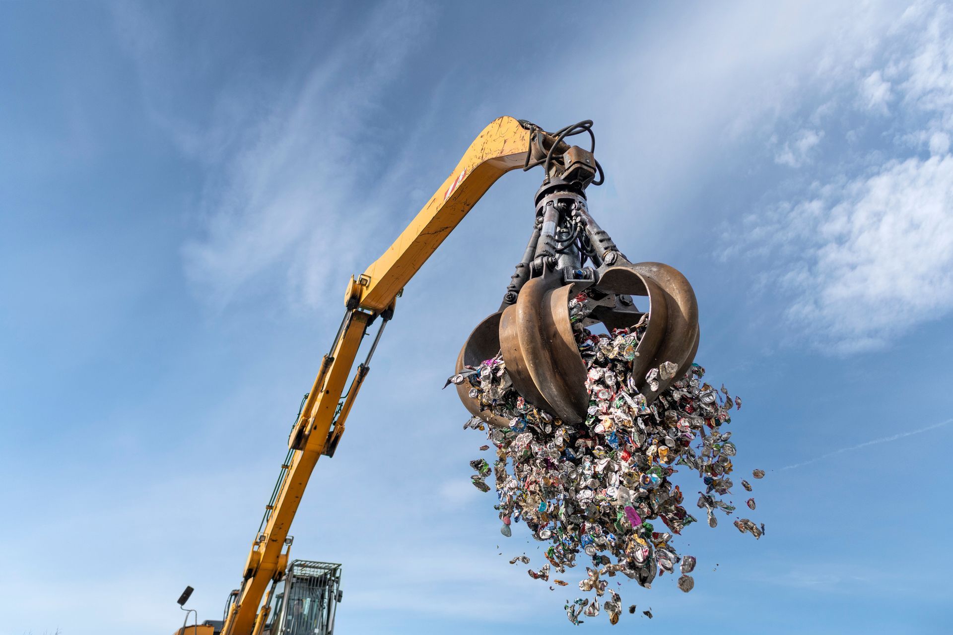 A yellow excavator lifting a clawful of mixed waste against a blue sky.