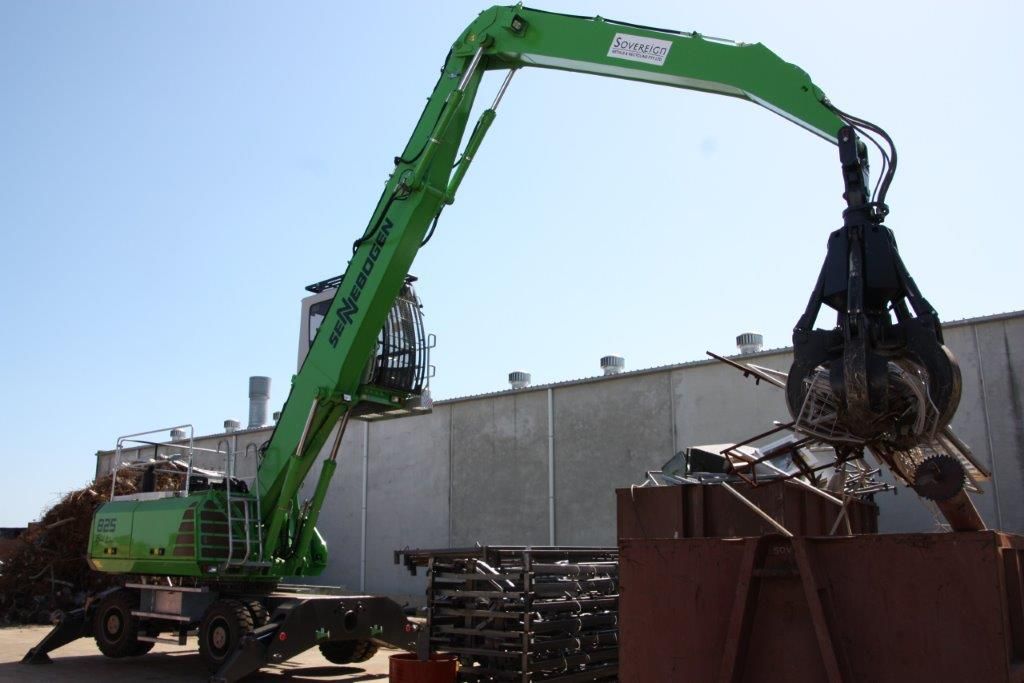 A green scrap metal handler, reaching over to grab metal pieces from a pile with its claw, set against a blue sky.