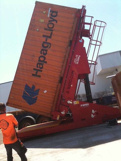 A red container lifting machine tilting an orange Hapag-Lloyd shipping container. A person in an orange shirt is nearby.