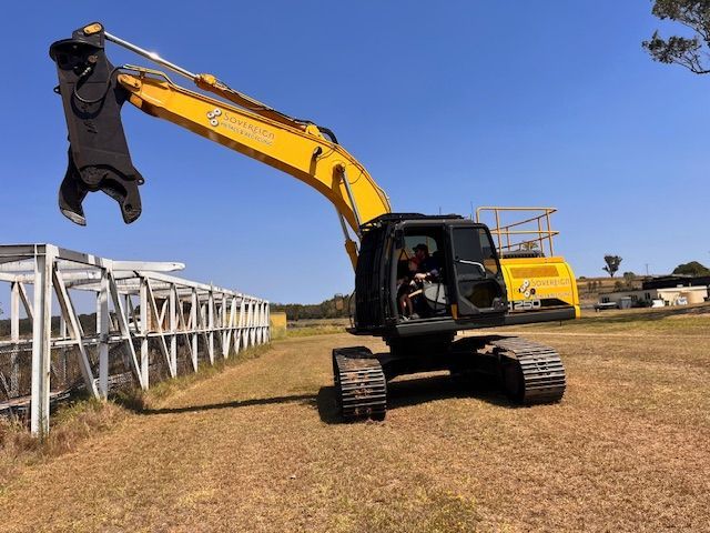 Yellow excavator with a grapple attachment on a grassy field under a clear blue sky. A person operates the machine.