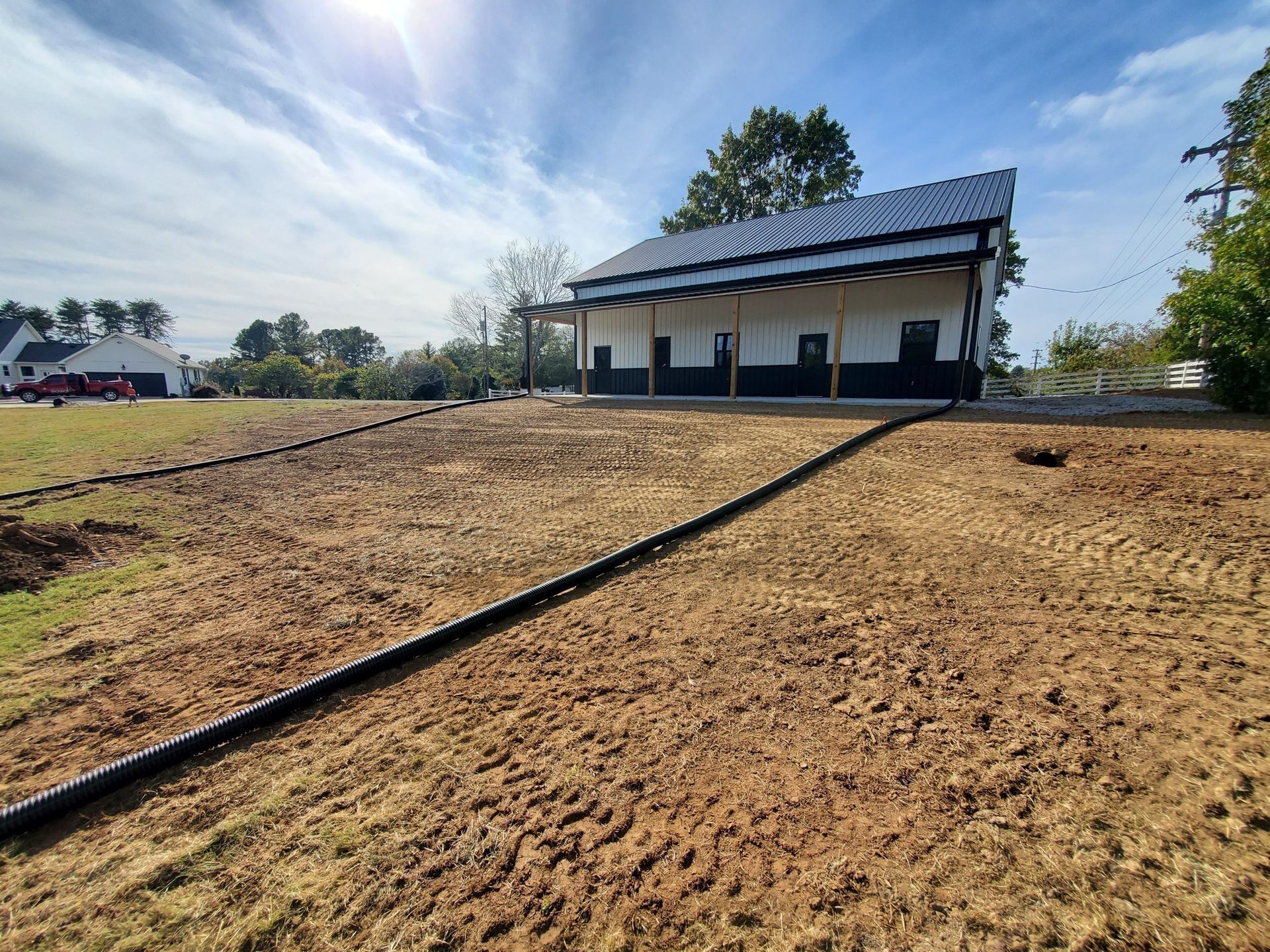 A modern house with a black roof and porch, viewed from a dirt yard with a black drainage pipe running across the ground.