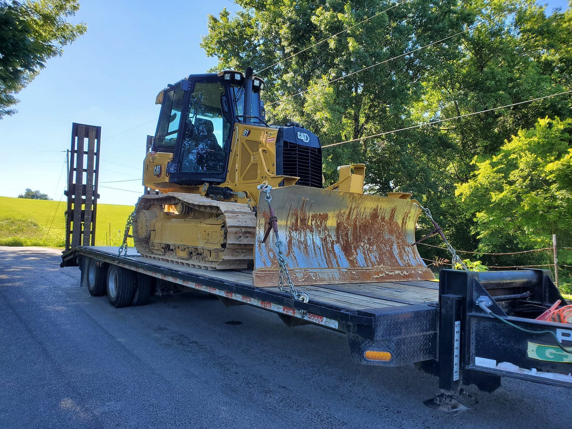 A yellow Caterpillar bulldozer sits parked on a flatbed trailer outdoors on a gravel surface near trees.