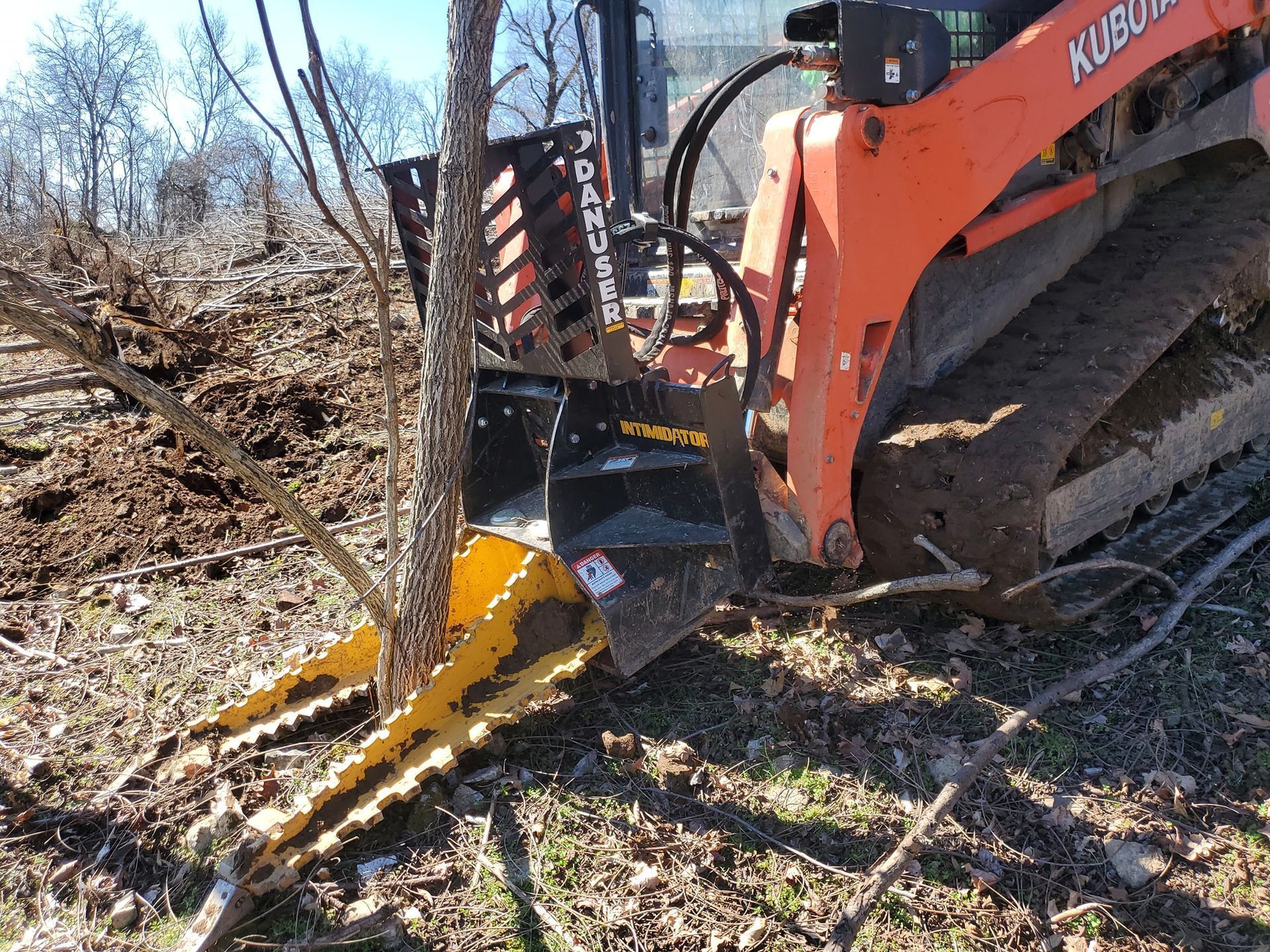 An orange skid-steer loader equipped with a yellow tree puller attachment gripping a tree trunk on a dirt field.