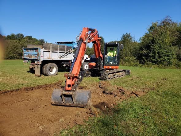 Orange excavator digging into a grassy field near a parked dump truck under a clear blue sky.
