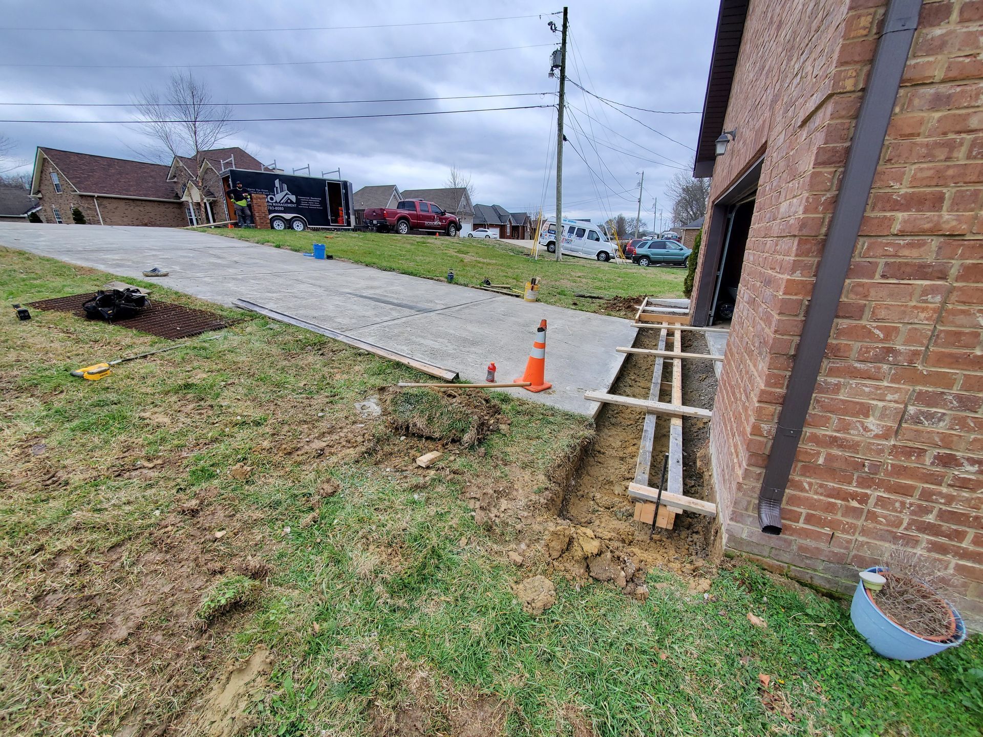 A construction trench with wood framing sits beside a brick house and concrete driveway under a cloudy sky.