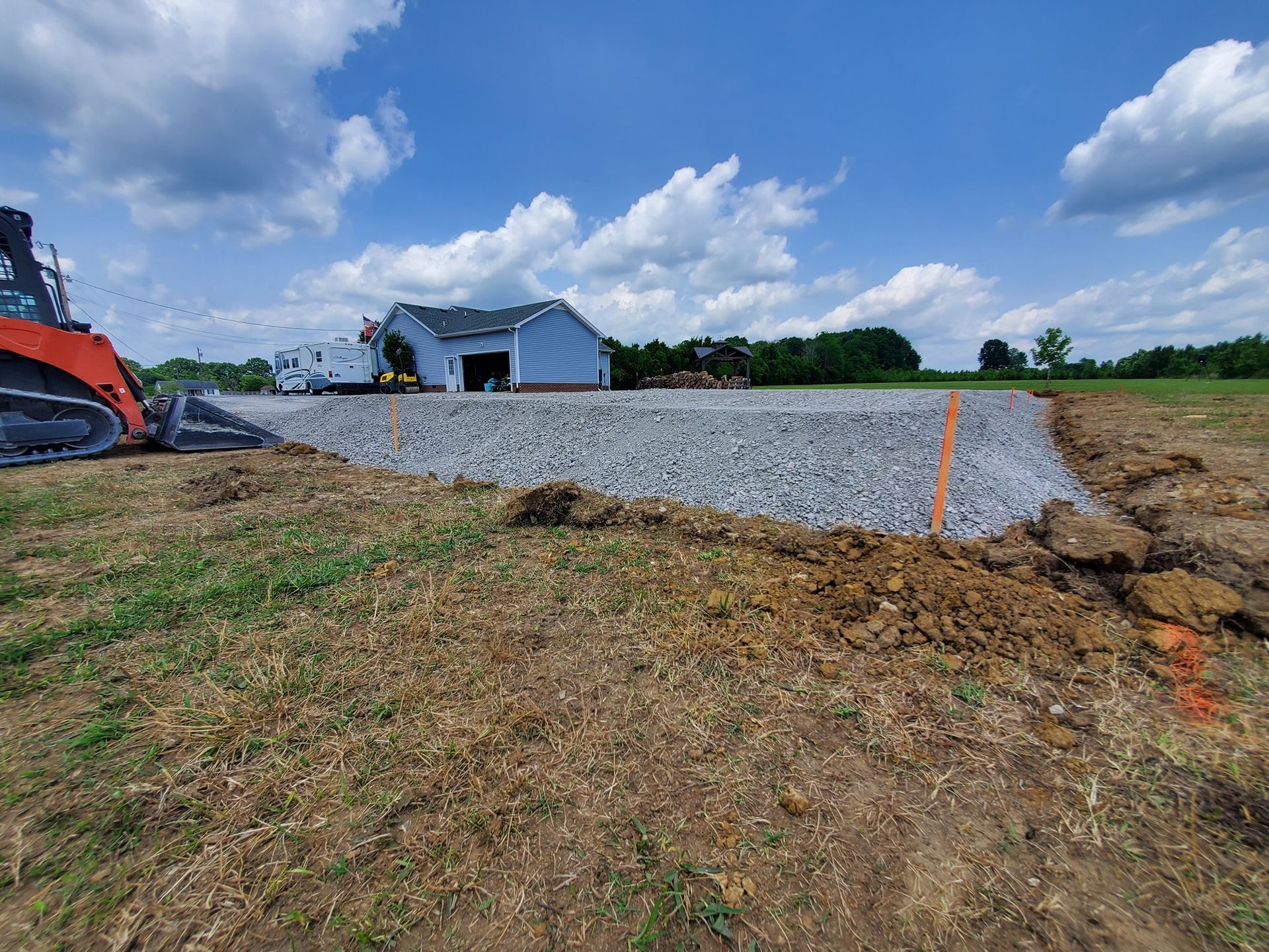 A construction site with a freshly graveled driveway, an orange piece of machinery, and a house in the distance.