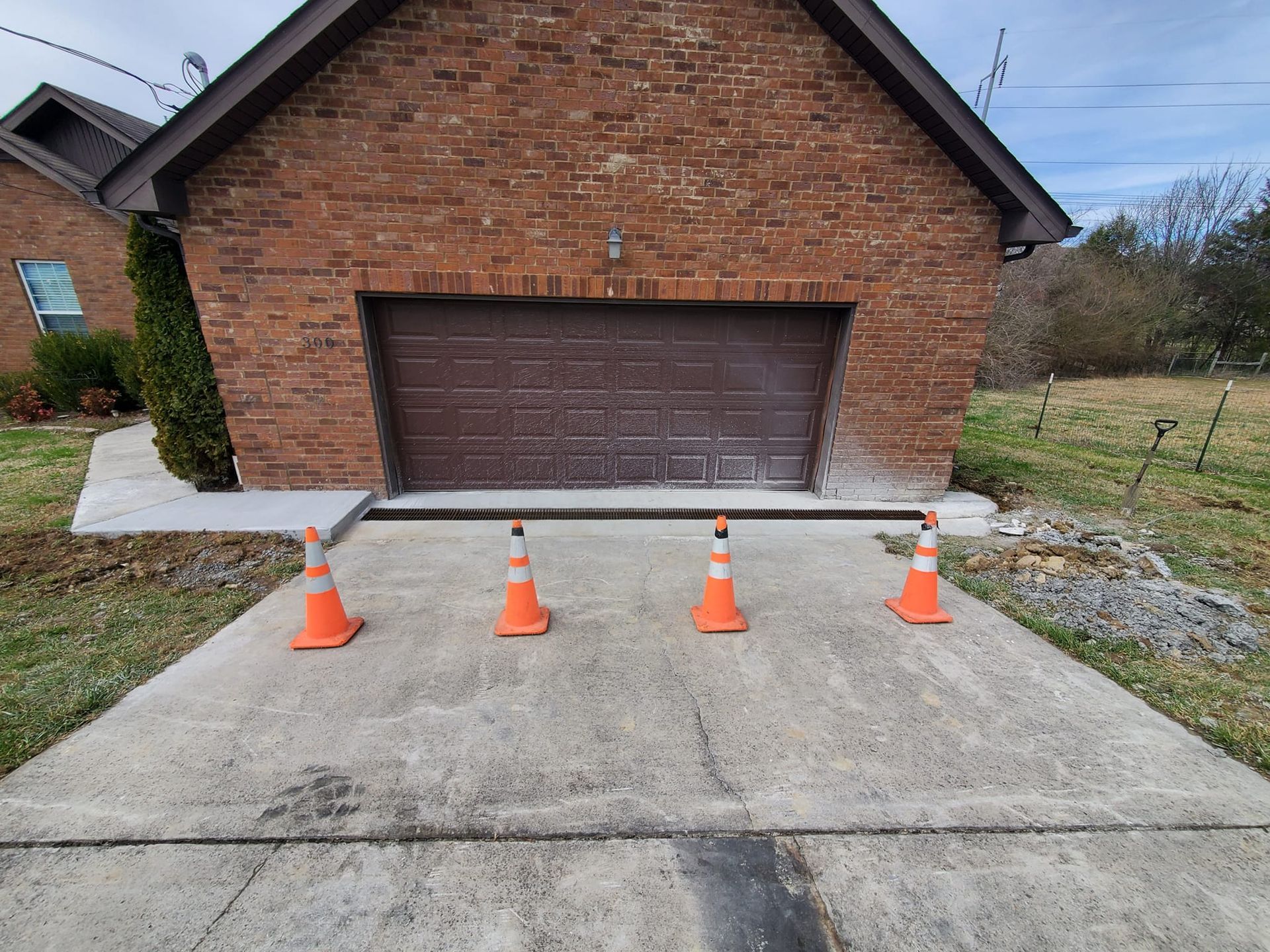 Four orange safety cones stand in a row before a gap in the concrete driveway in front of a brick garage.