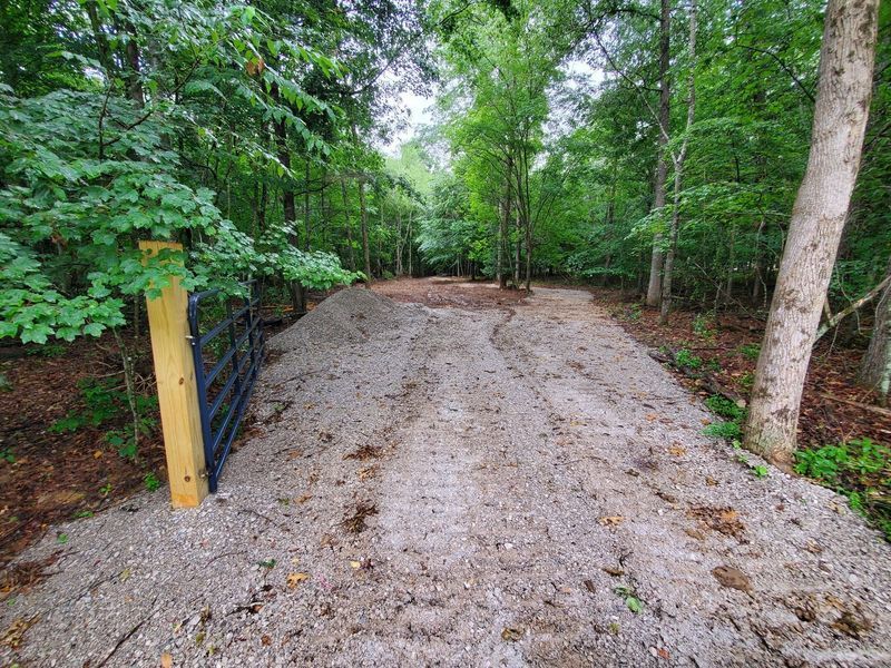 A gravel driveway leads into a wooded area, framed by a wooden post and a partially visible metal gate.