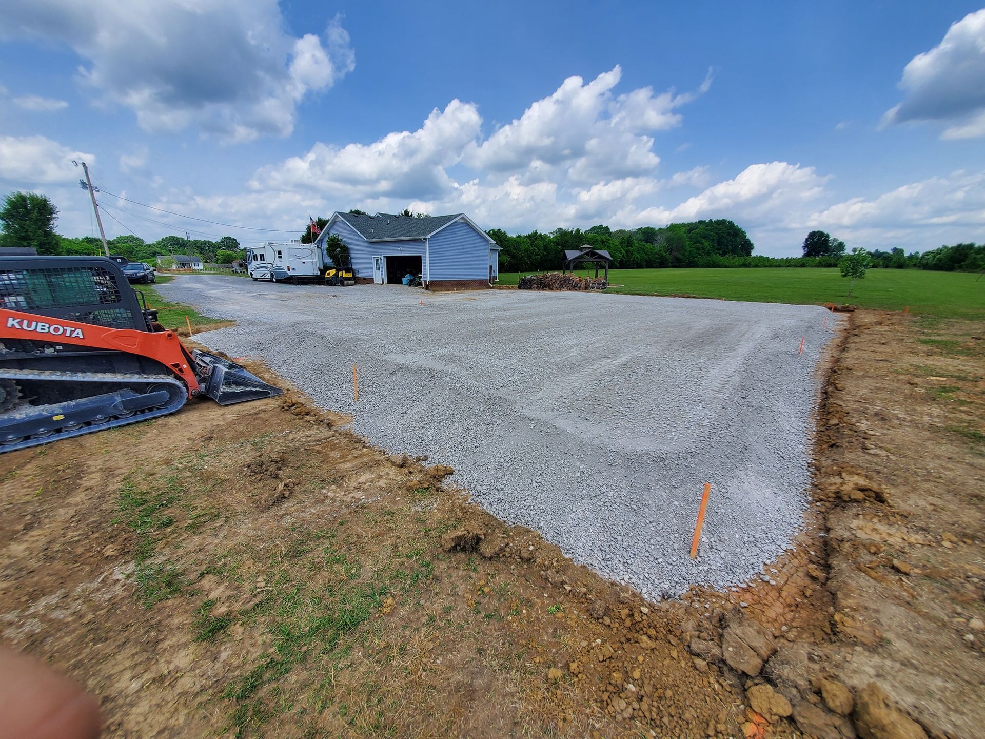 An orange skid steer sits next to a newly laid gravel driveway leading toward a house under a bright blue sky.