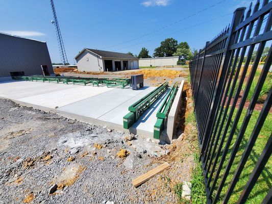 Construction site featuring a new concrete slab with green metal roller conveyors, bordered by a black fence on the right.