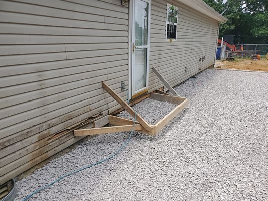 A partially constructed wooden frame for a small deck or landing sits outside a door on the side of a beige-sided house.