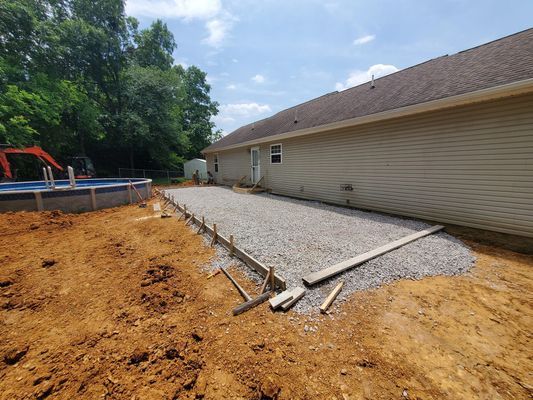 A gravel patio foundation framed with wooden boards alongside a house with tan siding and an adjacent swimming pool.