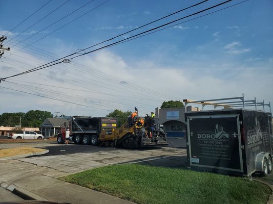 A construction crew uses a paver and dump truck to repave a parking lot on a sunny day.