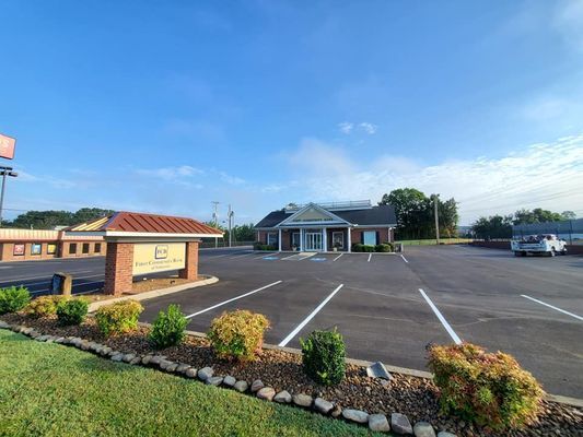 A brick building with a sign stands behind a paved parking lot under a blue sky, bordered by a landscaped grassy area.