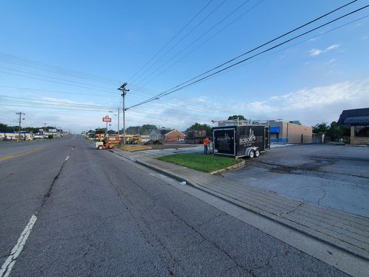 A person in an orange shirt works by a trailer parked on a roadside patch of grass near an asphalt parking lot.