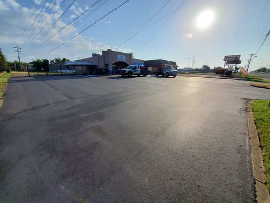 A bright, sunny parking lot with freshly paved black asphalt leading toward a commercial building with two parked trucks.