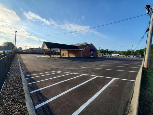A brick building with a drive-thru canopy sits beyond a freshly paved, marked parking lot under a clear blue sky.