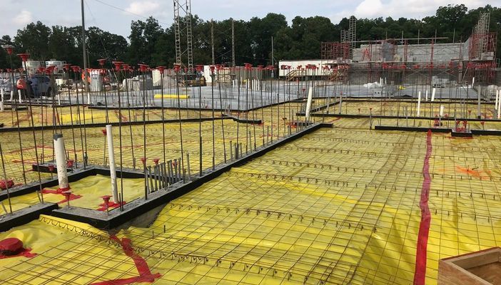 A construction site showing a concrete foundation prepared with yellow vapor barrier, wire mesh, and metal rebar columns.
