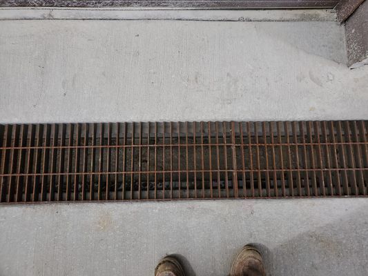 A metal grate covering a trench drain set into a concrete floor, viewed from above with a pair of boots visible below.