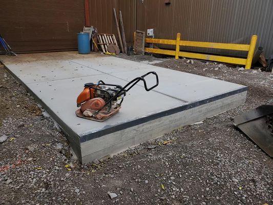 A plate compactor sits on a newly poured concrete pad in a gravel construction area next to a yellow railing.