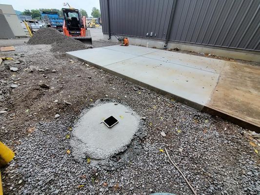 A newly poured concrete pad and a small circular concrete footing with a central access point in a gravel construction site.