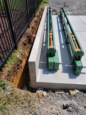 Two green industrial machinery tracks mounted on a concrete slab next to a metal fence and an excavated dirt trench.