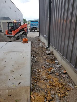 An orange skid steer loader sits near a concrete path next to a metal-sided building at a construction site.