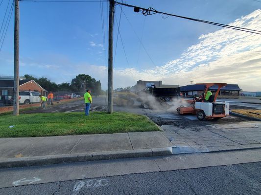 Construction workers use a small paving machine to repair a roadside area while others stand nearby on a grassy verge.