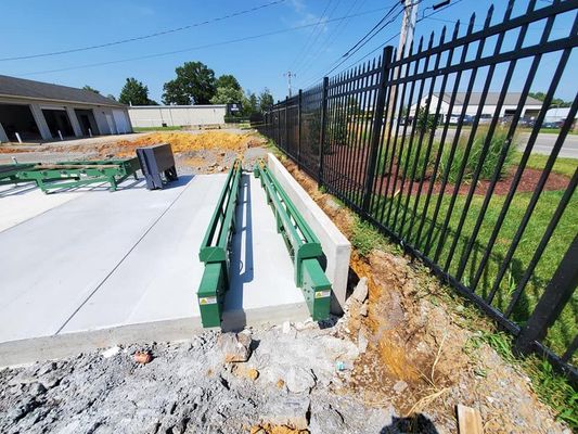Green industrial guide rails installed on a new concrete pad next to a black metal fence and an unpaved construction area.