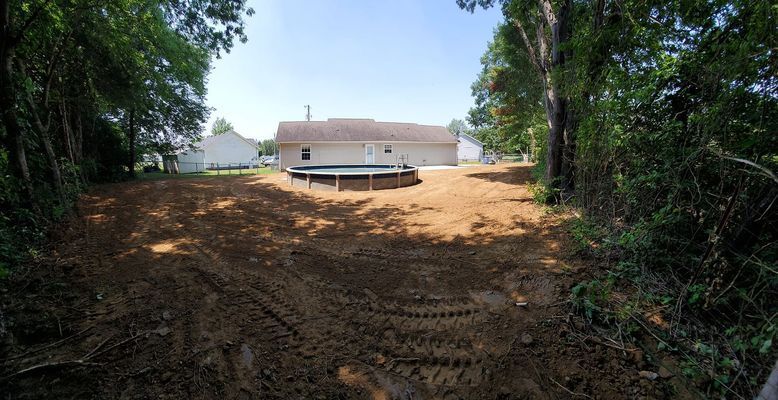 A wide, freshly graded brown dirt yard with a round above-ground pool behind a house, framed by trees on both sides.
