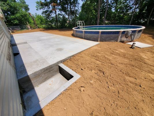 A concrete patio with a step down, adjacent to a house wall and an above-ground swimming pool in a dirt yard.