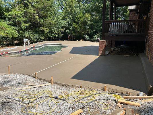 Newly poured concrete patio surrounding a backyard swimming pool next to a brick house with a wooden deck.
