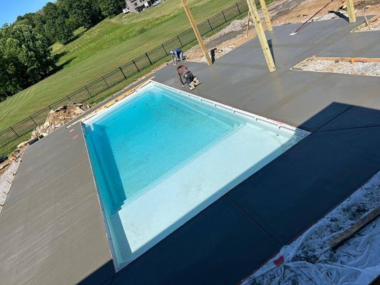 A high-angle view of a rectangular, light-blue swimming pool surrounded by a freshly poured, dark gray concrete deck.