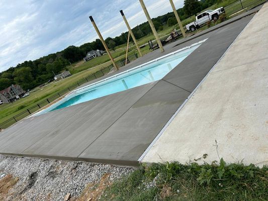 A freshly poured concrete pool deck borders a rectangular blue swimming pool set in a rural backyard landscape.