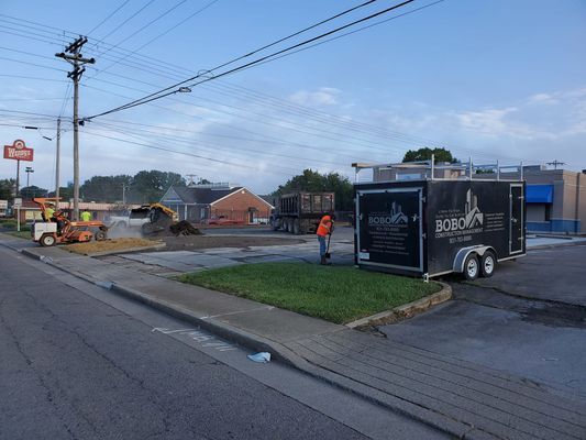 Construction crew working at an outdoor site with a black trailer parked on grass beside a paved street.