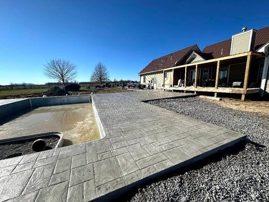 A concrete patio with a stone-like pattern surrounds a pool under construction next to a house with a wooden deck.