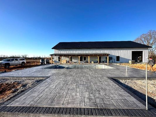 A long, white modern barn with a stamped concrete patio surrounding an empty in-ground pool under a clear blue sky.