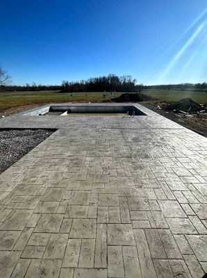 A wide view of a grey patterned stamped concrete patio surrounding an empty rectangular swimming pool in a field.